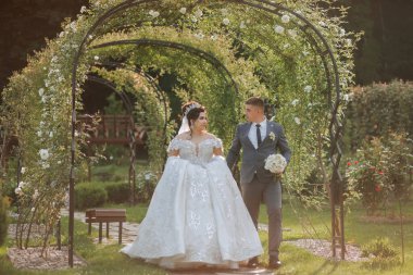 Gelin ve damat bahçede yürüyorlar, gelin beyaz bir elbise ve damat takım elbise giyiyor.
