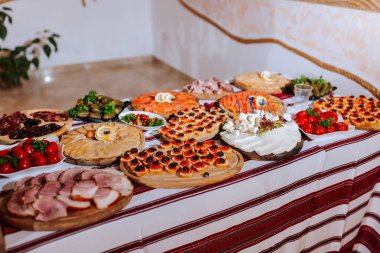 Bir masa pizza, sandviç ve meyve dahil olmak üzere çeşitli yiyeceklerle kaplıdır. Masa örtüsü kırmızı ve beyazdır ve arka planda birkaç saksı bitkisi vardır. Sahne bir şenlik sunar.