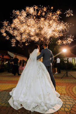 Bir gelin ve damat havai fişek gösterisinin önünde duruyor. Gelin beyaz bir elbise giyiyor ve damat bir takım elbise giyiyor. Çift diğer insanlarla çevrili.