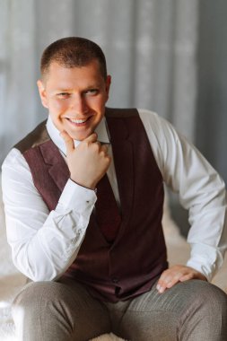 Takım elbiseli ve yelekli bir adam bir yatakta oturuyor. Gülümsüyor ve kameraya bakıyor.