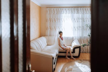 Oturma odasındaki kanepede oturan bir kadın. Beyaz bir elbise ve duvak takıyor.