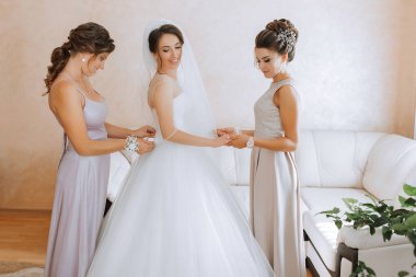 Üç kadın düğün için hazırlanıyor. Biri beyaz bir elbise giyiyor ve diğer ikisi de elbise giyiyor. Kanepenin ve saksı bitkisinin önünde duruyorlar.