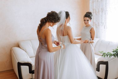 Üç kadın düğün için hazırlanıyor. Bir tanesi gelinin duvağını düzeltiyor. Diğer iki kadın onun arkasında duruyor, hazırlanmasına yardım ediyorlar.