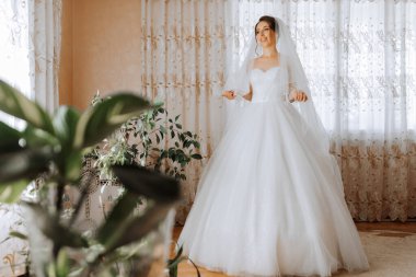 Beyaz gelinlikli bir kadın bir bitkinin önünde duruyor. Duvak takıyor ve mutlu.