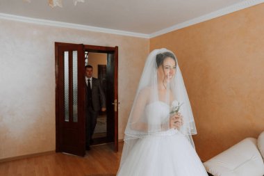 Bir gelin, elinde bir buket çiçekle bir adamın önünde duruyor. Adam takım elbise ve kravat takıyor. Gelin gülümsüyor ve kameraya bakıyor. Sahne mutlu ve kutlama dolu.