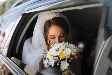 Bir gelin, elinde bir buket çiçekle arabanın içinde. Buketi tutuyor ve cep telefonuyla konuşuyor.