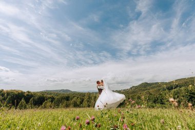 Bir gelin ve damat üzerlerinde güzel mavi bir gökyüzü olan bir tarlada duruyorlar. Gelin beyaz bir elbise giyiyor ve damat bir takım elbise giyiyor. Birbirlerini kucaklıyorlar.