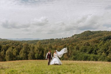 Bir gelin ve damat arka planda dağların güzel manzarasıyla bir tarlada yürüyorlar.