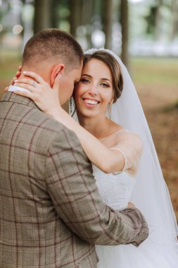 Bir gelin ve damat ormanda birbirlerine sarılıyorlar. Gelin beyaz bir elbise giyiyor ve damat da ekose bir takım giyiyor.
