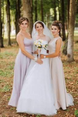 Üç kadın ormanda fotoğraf çektirmek için poz veriyor. Bir tanesi gelinlik giyiyor ve elinde buket tutuyor. Diğer iki kadın da elbise giyiyor ve gelinin yanında duruyorlar.