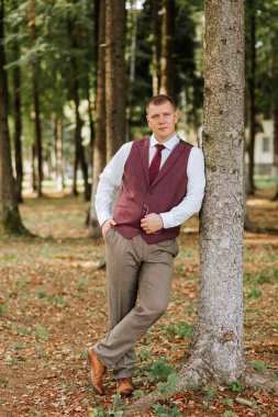 Kırmızı yelekli ve beyaz gömlekli bir adam bir ağacın yanında duruyor. Kahverengi pantolon ve kırmızı kravat takıyor.