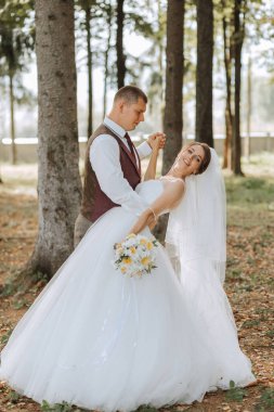 Bir gelin ve damat ormanda fotoğraf çektirmek için poz veriyorlar. Gelin elinde bir buket sarı çiçek tutuyor.