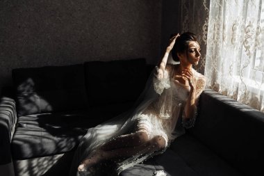 Beyaz elbiseli bir kadın pencereli bir odada kanepede oturuyor. Pencereden dışarı bakıyor ve düşüncelere dalmış.