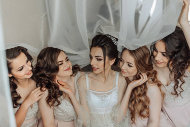 Beş kadından oluşan bir grup birlikte oturuyor ve biri gelinlik giyiyor. Kadınlar gülümsüyor ve birbirlerine eşlik etmekten zevk alıyor gibi görünüyorlar.