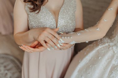 Gelinlikli bir kadın başka bir kadının elini tutuyor. Gelinlikli kadın uzun kollu bir elbise giyiyor ve elini diğer kadının koluna koyuyor.