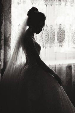 Gelinlikli bir kadın pencerenin önünde oturuyor. Elbise siyah beyaz ve kadın peçe takıyor. Sahne zarif ve romantik, çünkü kadın özel bir durum için giyinmiş.