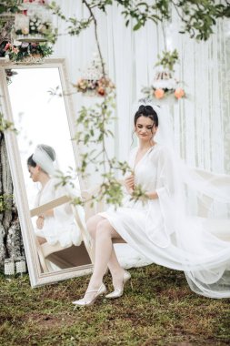 Beyaz elbiseli bir kadın aynanın önünde oturuyor. Bir duvak ve beyaz bir duvak takıyor.