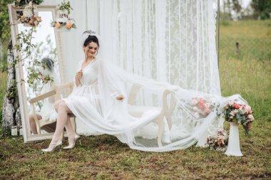 Beyaz peçeli ve beyaz elbiseli bir gelin bir sandalyede oturuyor. Etrafı çiçeklerle ve beyaz bir gölgelikle çevrili.