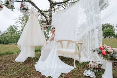 Beyaz bir elbise ve beyaz bir bankın önünde bir gelin poz veriyor. Sahne arka planda ağaç olan bir alanda geçiyor.