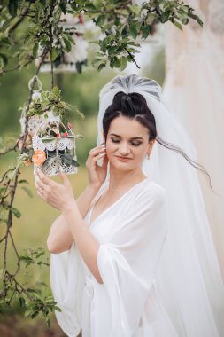 Beyaz bir elbise giyen bir kadın kuş kafesine bakıyor. Kadın peçe takıyor ve bir tarlada duruyor.