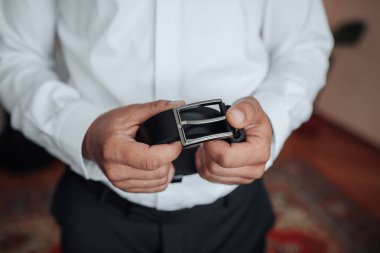 Bir adam elinde bir kemer tutuyor. Kemer siyah ve gümüş bir tokası var. Adam beyaz gömlek ve siyah pantolon giyiyor. Formalite ve zarafet kavramı