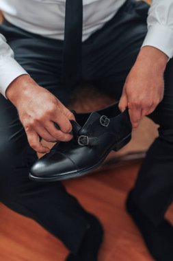 Bir adam ahşap bir zeminde oturuyor ve ayakkabılarını ayarlıyor. Ayakkabılar siyah ve önünde bir toka var. Adam beyaz gömlek ve siyah pantolon giyiyor.