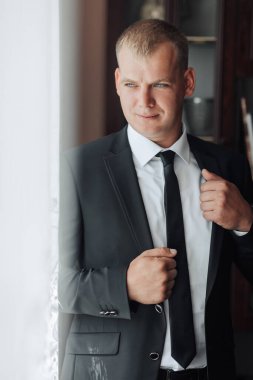 Takım elbiseli ve kravatlı bir adam pencerenin önünde kravatını düzeltiyor. Formalite ve profesyonellik kavramı. Çünkü adam takım elbise ve kravat giymiş.