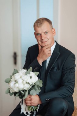 Takım elbiseli ve kravatlı bir adam kucağında bir buket çiçekle bir sandalyede oturuyor.