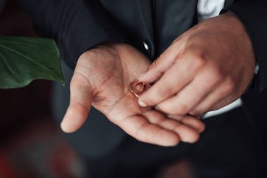 Bir adam elinde bir yüzük tutuyor. Yüzük altın ve kalp şeklinde. Adam takım elbise giyiyor ve kravat takıyor. Resmi ve zarif bir havası var.