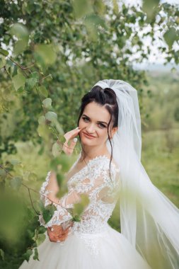 Beyaz gelinlikli bir kadın bir ağacın önünde poz veriyor. Gülümsüyor ve mutlu.