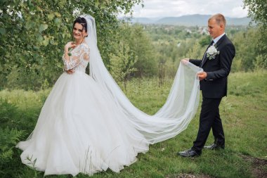 Beyaz peçeli bir adamla çimenli bir tepeden aşağı yürüyen bir gelin. Adam takım elbise ve kravat takıyor.