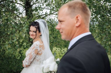 Bir gelin ve damat ormanda fotoğraf çektirmek için poz veriyorlar. Damat takım elbise giyerken gelin beyaz bir elbise ve peçe takıyor. Sahne romantik ve samimi.