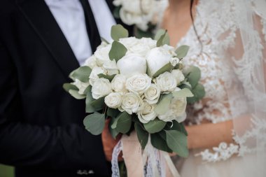 Bir gelin ve damat beyaz bir buket çiçek tutuyor. Gelin beyaz bir elbise giyiyor ve damat siyah bir takım giyiyor.