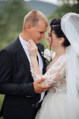 Bir gelin ve damat düğün fotoğrafında birbirlerine sarılıyorlar. Gelin beyaz bir elbise giyiyor ve damat siyah bir takım elbise giyiyor. Sahne mutlu ve romantik.