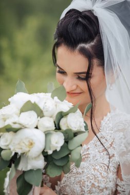 Bir gelin elinde bir buket beyaz çiçekle kameraya bakıyor. Gelin beyaz bir elbise ve duvak takıyor.