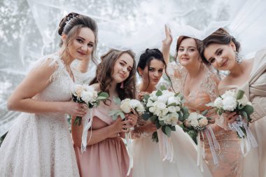 Bir grup kadın fotoğraf için poz veriyor, her biri bir buket çiçek tutuyor. Kadınlar düğün kıyafetleri giyiyorlar ve sahne açık havada çekiliyor. Sahne neşeli ve kutlayıcıdır.