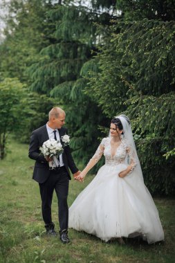 Bir gelin ve damat ormanda yürüyor, el ele tutuşuyorlar ve etrafları ağaçlarla çevrili. Gelin beyaz bir elbise giyiyor ve damat bir takım elbise giyiyor. Sahne romantik ve huzurlu.