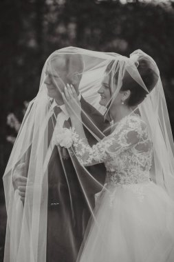 Bir gelin ve damat peçenin altında sarılıyorlar. Gelin beyaz bir elbise giyiyor ve damat bir takım elbise giyiyor. Sahne samimi ve romantik.