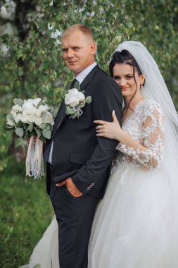 Bir gelin ve damat parkta fotoğraf için poz veriyorlar. Gelin beyaz bir elbise giyiyor ve damat bir takım elbise giyiyor. İkisi de elinde çiçek tutarken gelin elinde buket tutuyor.