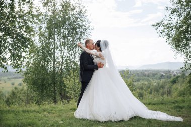 Bir gelin ve damat tarlada öpüşüyor. Gelin beyaz bir elbise giyiyor ve damat bir takım elbise giyiyor. Sahne huzurlu ve romantik.