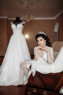 Genç bir kadın beyaz bir gelinliğin yanında bir kanepede oturuyor. Elbise sergileniyor ve kadın fotoğraf için poz veriyor. Zarafet ve kutlama kavramı