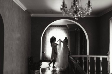 Bir kadın düğün için hazırlanıyor ve başka bir kadın ona yardım ediyor. Gelin beyaz bir elbise giyiyor ve diğer kadın da elbise askısı tutuyor. Sahne bir koridorda geçiyor.