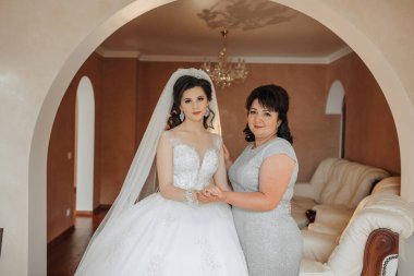 Bir gelin ve annesi bir kanepenin önünde duruyorlar. Gelin beyaz bir elbise ve duvak takıyor. Anne gelinin elini tutuyor. Sahne sıcak ve sevgi dolu.