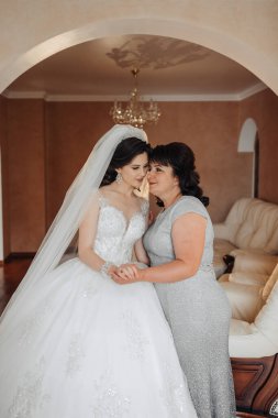 Bir gelin ve annesi oturma odasında sarılıyorlar. Gelin beyaz bir elbise giyiyor ve anne gri bir elbise giyiyor. Sahne sıcak ve sevgi dolu.
