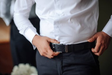 Beyaz gömlekli ve siyah kuşaklı bir adam pantolonunu düzeltiyor. Formalite ve detaylara dikkat etme kavramı. Adam kıyafetini düzgün bir şekilde giydiğinden emin oluyor.