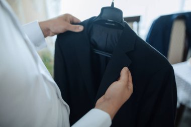 Bir adam elinde siyah bir ceket tutuyor ve ona bakıyor. Ceket askıda asılı.