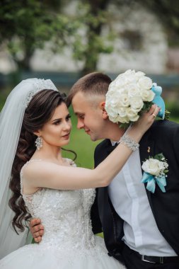 Bir gelin ve damat parkta birbirlerine sarılıyorlar. Gelin beyaz bir elbise giyiyor ve damat siyah bir takım elbise giyiyor. Gelin elinde bir buket beyaz gül tutuyor.