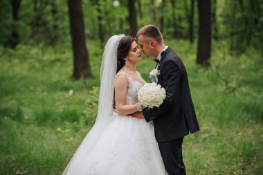 Gelin ve damat ormanda duruyorlar, gelin beyaz bir elbise giyiyor ve damat siyah bir takım elbise giyiyor. Ağaçlar ve otlarla çevrili el ele tutuşup öpüşüyorlar.