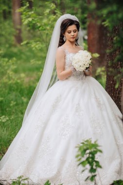 Elinde bir buket çiçek tutan beyaz elbiseli bir kadın. Kadın ağaçlarla çevrili bir çim tarlasında duruyor. Huzurlu ve huzurlu bir sahne.