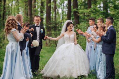 Bir gelin ve damat düğün partileriyle ormanda yürüyorlar. Gelin beyaz bir elbise giyiyor ve damat bir takım elbise giyiyor. Düğün ekibi alkışlıyor.
